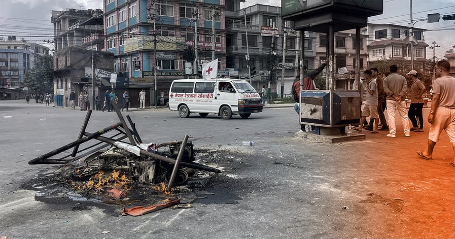 Street burning with people watching, ambulance in background Description: Protesters set fire to debris and barricades in the middle of a road in Nepal during the September 9 protest. Locals and bystanders watch as flames burn, while an ambulance passes, symbolizing the tension and emergency state in the city.